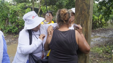 En kvinna får vaccin i armen utomhus under ett paraply av en kvinna i vitt och Läkare Utan Gränsers logga på hatten.