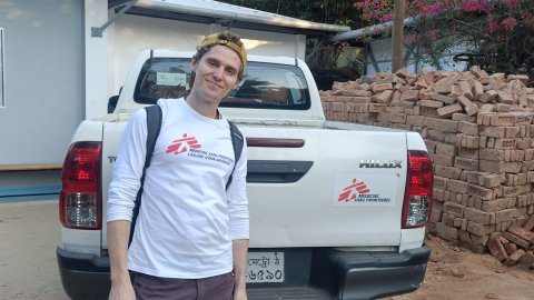 En man med keps och vit Läkare Utan Gränser-tröja utomhus framför en bil.