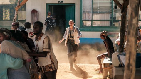 Kvinna i läkarväst srpinger ut ur ett hus och in i en folksamling. Stillbild från filmen Före mörkret