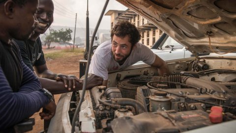 En man mekar med en bilmotor samtidigt som han pratar med två skrattande kollegor.