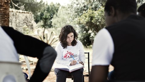 En kvinna och några män sitter på stolar i en ring under ett träd. Kvinnan antecknar i en bok, männen har ryggen vänd mot kameran.