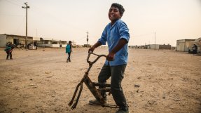 Haussian, en 13-årig pojke, står leende med sin trasiga cykel på en grusplan.