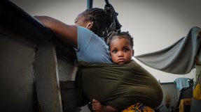 En mamma med ett litet barn i en sjal på ryggen lutar sig mot relingen på en båt och tittar ut över havet