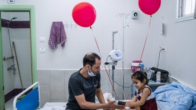 Mohammed Aboud sitter brevid sin fyra år gamla dotter Hala som ligger i en säng på barnavdelningen på al-Awda sjukhus i norra Gaza, Palestina. De tittar på en mobiltelefon och två röda ballonger hänger på sängen.