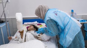 En sjuksköterska böjer sig över fyraåriga Halas säng på al-Awda sjukhus i norra Gaza, Palestina, för att trösta henne. Halas högra fot är gipsad.