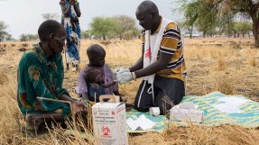 En hälsoinformatör från Läkare Utan Gränser står på knä och testar ett litet barn för malaria utomhus under ett träd i Thaker, Sydsudan. Barnet sitter i en vuxens knä på en filt, bredvid sitter ytterligare en vuxen.