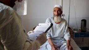 En äldre man sitter i en sjukhussäng och pratar med en medarbetare.