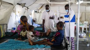 Bria sitter på sängen tillsammans med sina tre barn på sjukhuset i Tukra, Tchad. Bredvid står två läkare.
