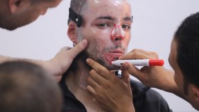 En man har en plastmask för ansiktet. En annan personer målar på masken med en röd penna.