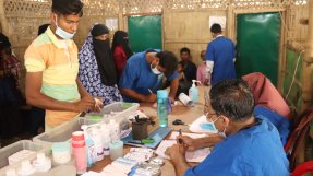 En man står vid ett bord vid vilket en blåklädd man sitter med mediciner framför sig.