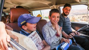 Två pojkar och en man sitter i framsätet av en bil, en pojke vinkar mot kameran och ler