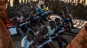 en grupp skolelever med skolböcker sitter i två rader på marken i en hydda