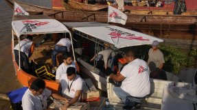 Flera personer sitter på träbåtar och lassar kartonger av och på, Läkare Utan Gränsers logotyp syns på flera ställen.