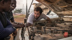 En man mekar med en bilmotor samtidigt som han pratar med två skrattande kollegor.