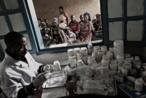 En läkare sitter vid en stor mängd mediciner. Människor väntar utanför ett fönster.