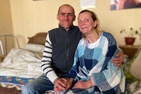 Sergej och Natasha sitter på en säng och håller varandras händer. 