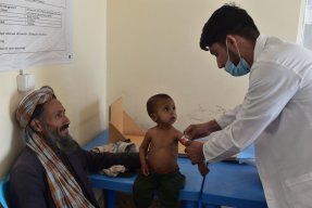 En sjuksköterska undersöker ett barn som sitter på en brits på Läkare Utan Gränsers klinik i Herat. På en stol bredvid sitter en äldre man och ler mot barnet och sjuksköterskan.