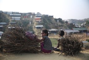 En flicka i flyktinglägret Cox's Bazar, Bangladesh, sitter på huk bredvid en pojke och en hög med grenar och tittar mot kameran.
