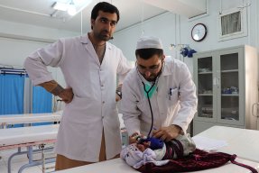 Två män iklädda vita sjukhusrockar står vid en brits på Boost sjukhus, Lashkar Gah, Afghanistan. På britsen ligger en liten bebis.