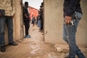 Människor står vid en dörröppning i förvaret Dahr-el-Jebel i Libyen. Det är lerigt på marken.