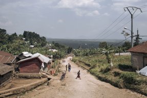 Personer går längs en slutande grusväg. På sidorna syns enklare hus i trä. 