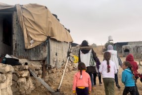Två personer i Läkare Utan Gränser västar går längs en gatan. Bakom dem går en grupp barn. Husen runt omkring är byggda i betong med tältdukar som tak.