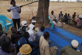 En grupp män sitter i en cirkel. En kvinna hållandes en mikrofon står bredvid.