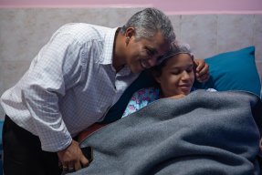 En man håller om en kvinna som ligger i en sjukhussäng. Tillsammans tittar de på ett nyfött barn.