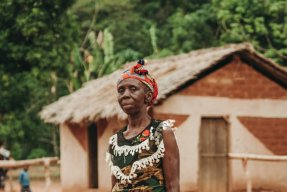 En medelålderskvina står med en neutral blick tittandes mot kameran, i bakgrunden syns ett tegelhus i ett stepplandskap.