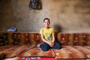 En leende pojke i 10-årsåldern med gul t-shirt sitter på en madrass på golvet i ett kalt rum