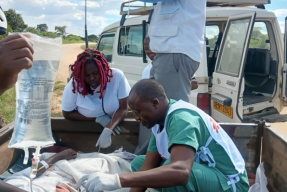 Tre personer i sjukhuskläder ger dropp till en person som ligger ner i någon form av fordon.