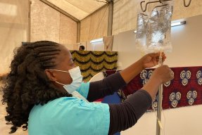 En kvinna i gröna sjukhuskläder och munskydd fixar till en påse dropp.