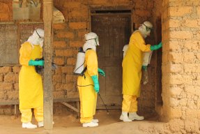 Tre personer i gula dräkter, handskar och masker står vid en dörr till ett teglat hus.