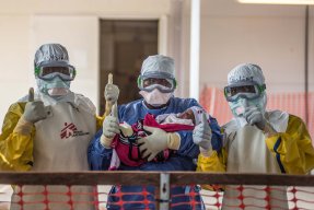 Tre vårdpersonal i skyddsdräkter från vårt ett av våra ebolacentrum gör tummen upp med ett spädbarn i famnen