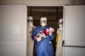 Utrustade läkare kollar in i kameran där en av tre har ett barn i famnen.