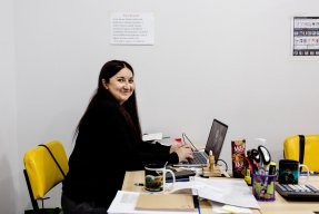 Kvinna sitter vid datorn och tittar in mot kameran och ler.