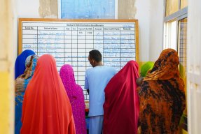 Kvinnor i färgglada kläder och en man i blått vända mot en whiteboard.