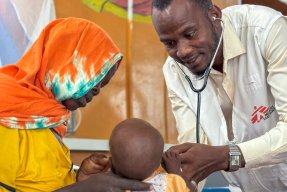En man med stetoskop och Läkare Utan Gränser-väst undersöker ett barn. En kvinna är med barnet.