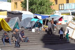 Tältläger i urban miljö. Två personer i Läkare Utan Gränser-västar pratar med en grupp migranter