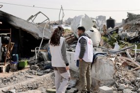 En man och en kvinna från Läkare Utan Gränser står framför rasmassorna av en byggnad som sprängts.