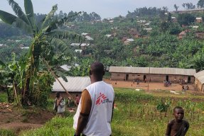 En man i Läkare Utan Gränser-väst med ryggen vänd mot kameran vänd mot gröna kullar, bananplantor och hus i bakgrunden.