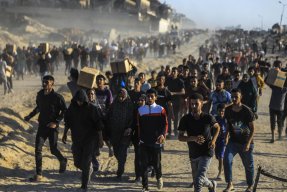 Människor i en stor folkmassa rör sig längs en dammig väg, flera bär kartonger