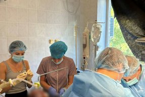 Fyra personer iklädda munskydd varav två blå sjukhusrockar i ett rum på ett sjukhus.