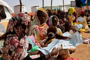 Kvinnor och barn sitter i skugga utanför klinik