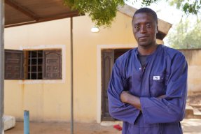 En man i blå overall står utomhus med armarna i kors och tittar in i kameran.