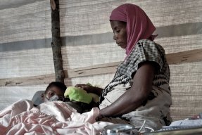 En kvinna med sjal sitter på en enkel säng med ett barn liggandes bredvid henne. Barnet bär en syrgasmask.