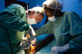Två läkare i operationsrockar och munskydd syr ihop ett sår på en fot.