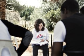 En kvinna och några män sitter på stolar i en ring under ett träd. Kvinnan antecknar i en bok, männen har ryggen vänd mot kameran.