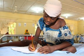 Flicka med sjukdomen noma efter operation sitter i en säng och ritar