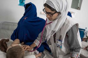 En kvinnlig läkare lyssnar på ett barns andning på ett sjukhus i Afghanistan.Bredvid på sängen sitter en anhörig kvinna med ansiktet täckt av en slöja. 